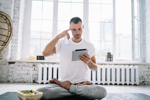 Concentrated young man in sportswear sitting on rubber mat with legs crossed and resting with tablet while meditating in bright studio