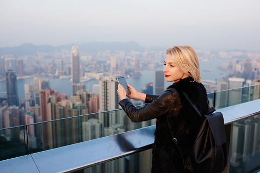 Side view of content female traveler in casual clothes with backpack standing on rooftop of contemporary building in Hong Kong and browsing social media on mobile phone and looking away