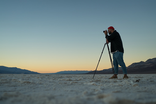 From below of male wearing black shirt and jeans shooting photo on photo camera with tripod while standing on salt pans of Badwater Basin in Death Valley in USA