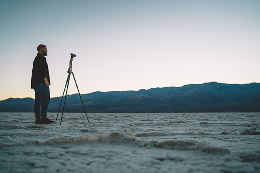 Side view of full body young male traveler standing near tripod and shooting landscape of dry mountainous desert at sunset