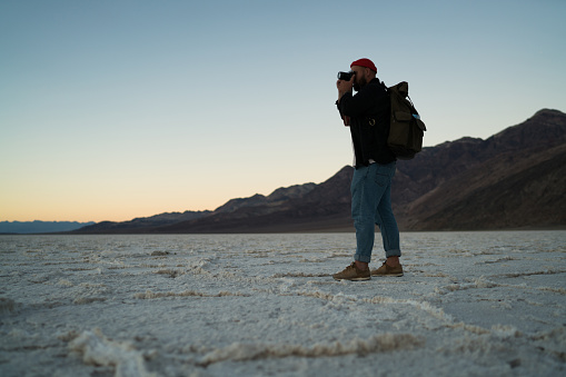 Side view of full length of male traveler in outwear with backpack and hat taking photos of Badwater Basin with rocky mountains in twilight