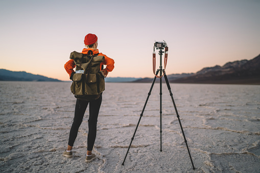 Back view of full length of female photographer looking at natural landscape of Death Valley in America while taking picture of Badwater Basin with photo camera on tripod