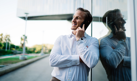 Cheerful businessman in white shirt smiling near urban building enjoying positive international conversation, happy male entrepreneur calling to friendly partner using roaming wireless connection