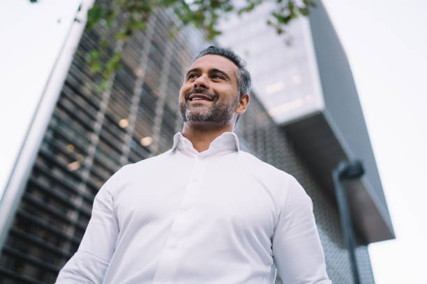 a continuación, vista del empresario de 40 años vestido con una camisa blanca formal mirando hacia otro lado y sonriendo, exitoso hombre de negocios de mediana edad disfrutando de un descanso laboral en el distrito financiero del centro de la ciudad - 35 40 years fotografías e imágenes de stock