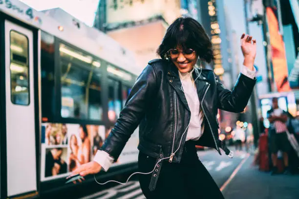 Photo of Carefree woman in fashionable clothing listening funny audio and dancing during evening walk in New York City, cheerful female enjoying adolescent music podcast during nightlife lifestyle in Manhattan