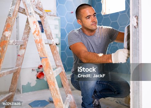 istock The technology of laying tile, repair and decoration. Focused man puts porcelain tiles on wall sitting squat at bathroom 1432431635