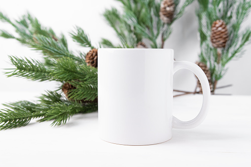 White ceramic cup with space for text or logo on a wooden table with Christmas tree branches in the background