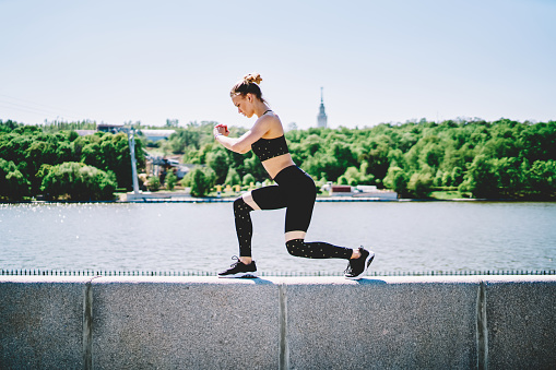 Side view of sporty woman in activewear exercising and doing lunges while showing off strong body on background with river and forest