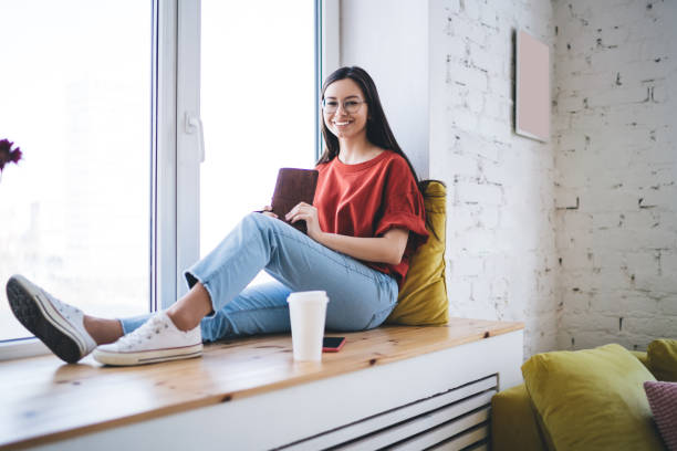 istock Smiling woman sitting on window sill and looking at camera 1432437833