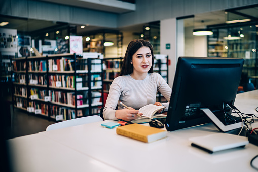 Young confident smiling woman in casual clothes reading textbook and browsing computer while sitting at table in modern library