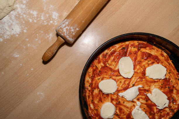 Kitchen table with dough, rolling pin and an homemade unbaked pizza with tomato sauce and mozzarella