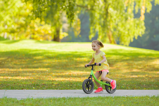 Happy beautiful little girl fast running and riding on first bike without pedals on sidewalk at city park in warm summer day. Cute 3 years old toddler. Side view. Learning to keep balance.