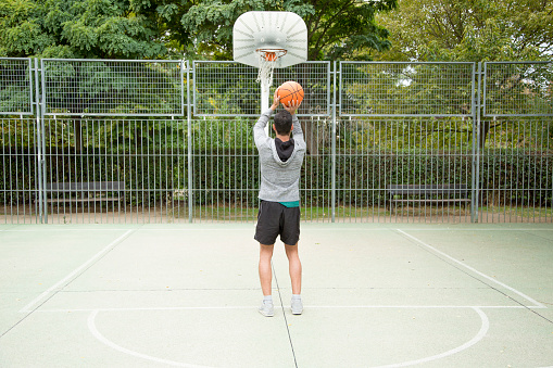 Rear view of a sportive man throwing a ball from the three-point line on an outdoor basketball court