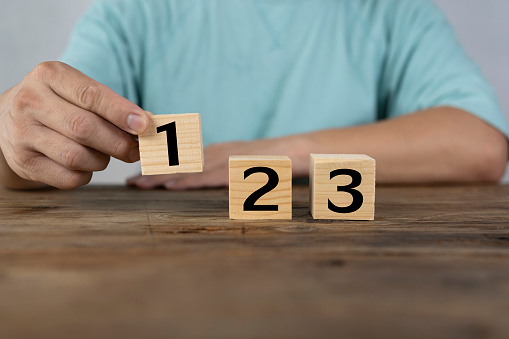 Close-up of a man holding a wooden block numbered one, two and three on a wooden table.