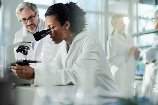 Black female scientist looking through a microscope while her colleagues are around her.