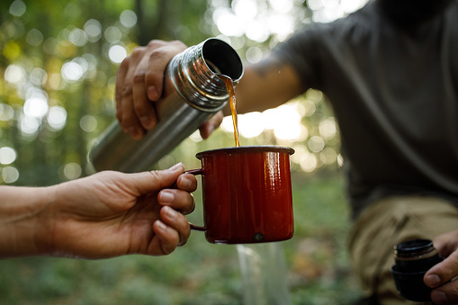 Close up shot of unrecognizable woman holding a mug while her male friend is pouring coffee from thermos while sitting in the forest, taking a break from hiking.