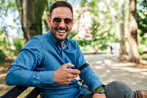Picture of happy adult man with sunglasses, sitting in the park on the bench, using his phone.
