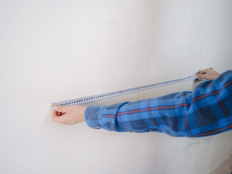 Side view of serious man using measuring tape in room with underfloor heating pipes. Designer in shirt taking measurements while working on renovation of apartment.