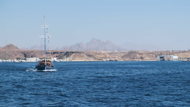 An old wooden ship, bow view, with lowered sails, emerges from the bay. View of the harbor in Sharm El Sheikh, desert, sand, pier.