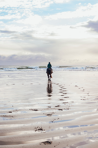 Rear view of a woman horseback riding along the beach in the North East of England. The scene is tranquil and calm. The horse is leaving footprints in the sand.