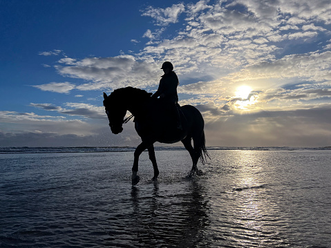 Low angle wide side shot of a woman horseback riding along the beach in the North East of England. The scene is tranquil and calm and the sky is dramatic behind her at sunrise.