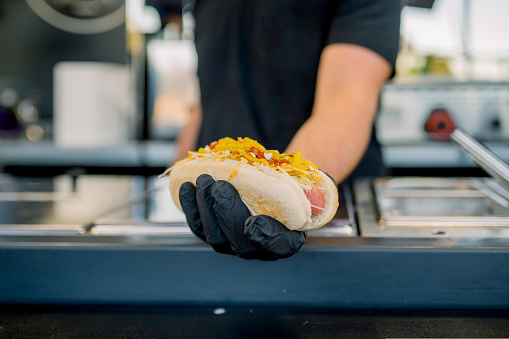 Faceless vendor standing at counter and stretching hand in glove with prepared hot dog to camera in selective focus