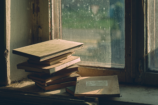 Somewhere in an abandoned house, old dusty books and ledgers are lying on a dirty windowsill, the light of the evening sun is coming through the dirty window, an atmospheric still life of abandoned things