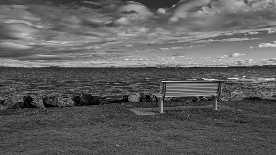 A grayscale shot of a wooden bench at the lake shore under a cloudy sky