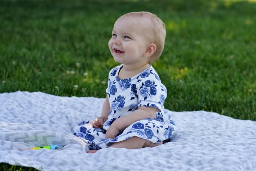 A baby girl wearing a white dress with blue flowers, sitting on the grass and expressing joy