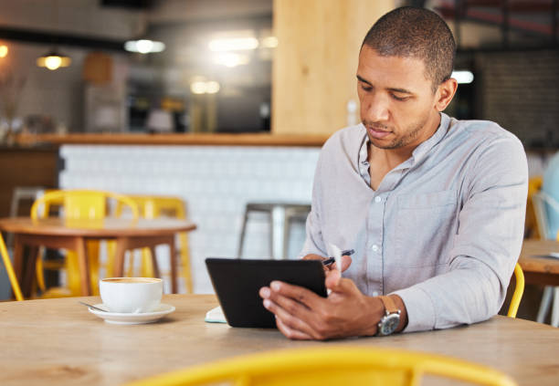 istock Serious freelance business man working in a coffee shop doing remote work for his startup. Young male entrepreneur using a tablet planning growth strategy in a cafe and writing notes 1432500021