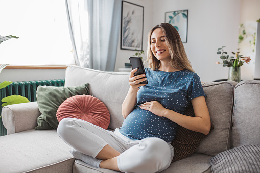 Young pregnant woman at home. She is sitting on sofa and using smart phone for online shopping or to pay bills.