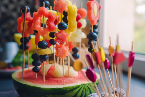 bunter fruchtdessert zum geburtstag eines kindes. verschiedene formen von früchten werden auf zahnstocher gelegt. zahnstocher stecken in einer halben wassermelone. wassermelone, ananas, melone und blaubeeren. - fruit salad fruit healthy eating making stock-fotos und bilder