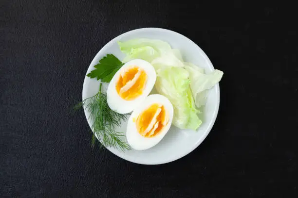 Halves of a hard boiled chicken egg with lettuce, dill and parsley on a plate on a dark background - top view