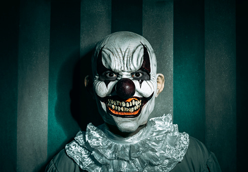 a creepy bald evil clown, in a gray costume with a white ruff around his neck, in front of a dirty and old circus tent, stares at the observer with a frightening smile