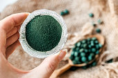 Female hand holds spirulina powder on a background of burlap and a spoon with green algae tablets