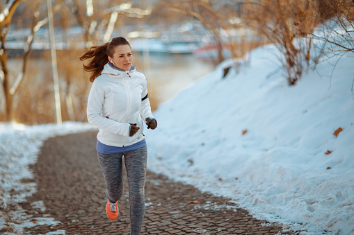 fit woman in white jacket running outside in the city park in winter.