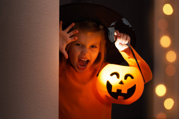 istock Halloween. Girl in witch hat hides and scares from corner with lantern pumpkin 1432531994