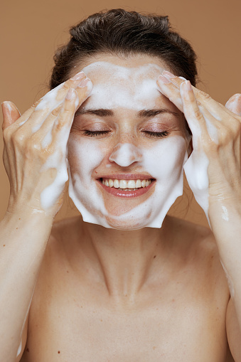happy middle aged woman with foaming facial cleanser washing face isolated on beige background.