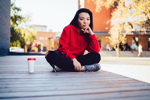 Portrait of attractive millennial woman sitting in lotus pose at urban setting and looking at camera during leisure time, beautiful Chinese woman dressed in stylish streetwear posing in city