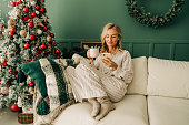 A woman is drinking coffee on Christmas morning and texting on her phone while sitting on the couch