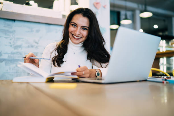 istock Portrait of cheerful female student 20s looking at camera during time for laptop e learning, happy Caucasian woman with education textbook rejoicing at desktop with modern netbook technology 1432552799
