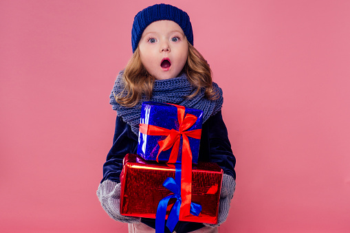 little blonde curly hairstyle girl in knitted pink hat ,scarf and mittens mountain of gifts christmas box pink background studio. new Year present in hands of a female child making a wish copyspace