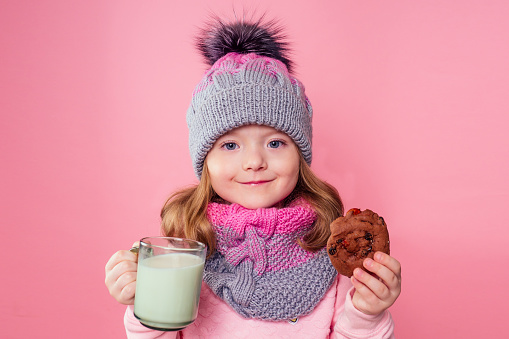 beautiful little girl drinks milk and eats cookies in a knitted hat and scarf on a pink background in the studio,christmas night.