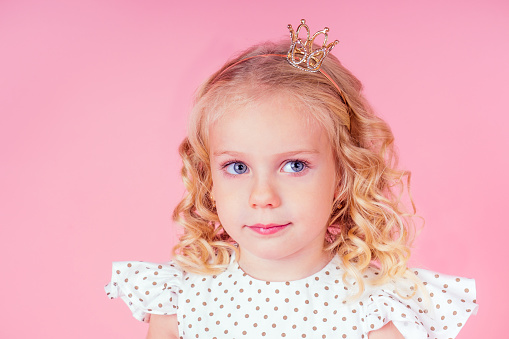 little girl beauty queen blue eyes, curls blonde hairstyle with a tiara crown on her head in a cute white dress in peas posing in the studio on a pink background.birthday celebration,Beauty contest