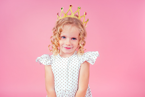little girl beauty queen blue eyes, curls blonde hairstyle with a tiara crown on her head in a cute white dress in peas posing in the studio on a pink background.birthday celebration,Beauty contest