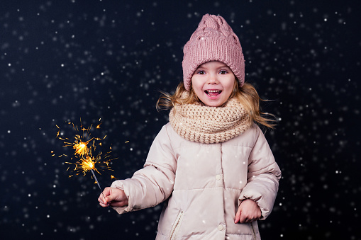 charming little girl in a knitted pink hat holding fireworks on black background in a studio.Cute blonde child with xmas dream.Happy kid enjoy the fire sparks. new year holidays eve of Christmas wish.