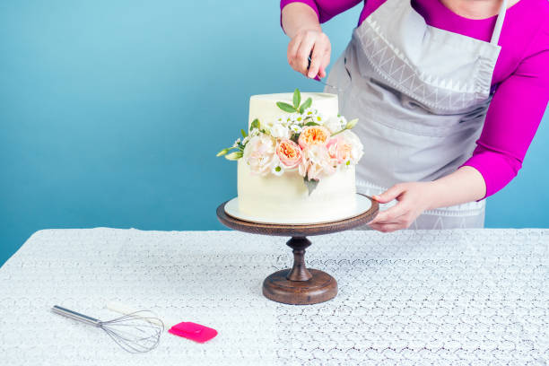 女性菓子職人(ペストリークック)は、青い背景にスタジオのレースのテーブルクロスでテーブルの上に新鮮な花で飾られた食欲をそそるクリーミーな白い2層のウェディングケーキを飾る - cake women confectioner photography ストックフォトと画像