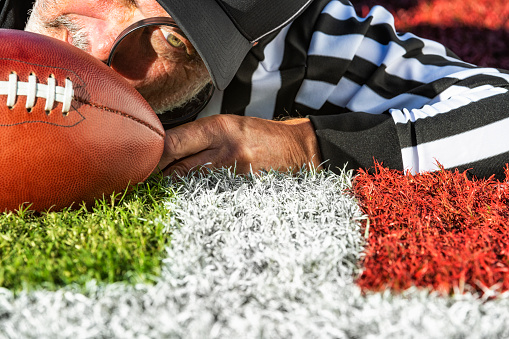 A close-up of an American football referee with a magnifying glass, checking to see if the ball “broke the plane” of the goal line. He is lying on the field.