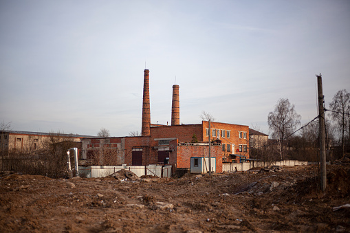 This is an image of the ruins of an old factory building. The building is made of red bricks and has a tall chimney. The factory is surrounded by overgrown vegetation. The sky is clear and blue.