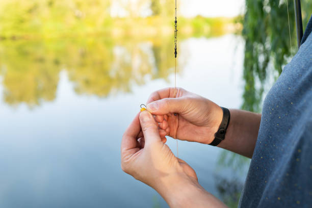 el concepto de cebo para peces. primer plano de la mano de un pescador encordando el cebo en una caña de pescar contra un lago azul. - lure loc fotografías e imágenes de stock